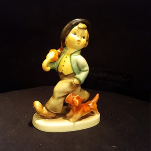 May include: A porcelain figurine of a boy in a blue jacket and brown pants walking with a brown and white dog. The boy is holding an umbrella over his shoulder.
