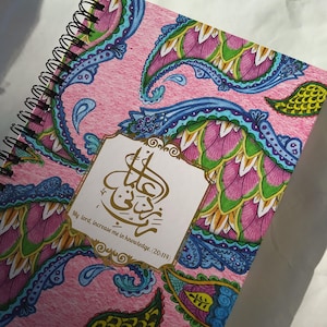 Pode incluir: Um caderno espiral rosa com um padrão paisley colorido e letras douradas. O texto diz "Meu Senhor, aumenta meu conhecimento. (20:114)"