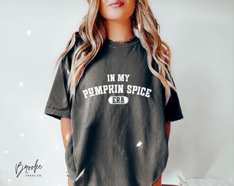 May include: Dark gray t-shirt with the text "IN MY PUMPKIN SPICE ERA" in white, with a white oval below the text. The shirt is a crew neck and has short sleeves. The shirt is worn by a person with long blonde hair.