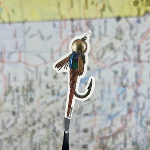 May include: A sticker of a fishing lure with a gold-coloured head, brown and blue feathers, and a large hook. The lure is attached to a dark grey handle. The background is a map.