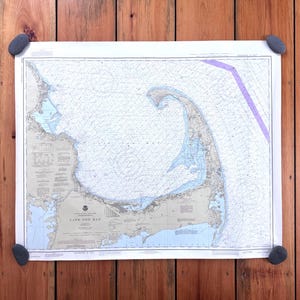 May include: A nautical chart of Cape Cod Bay, featuring detailed coastlines, depths, and navigational information. The map is secured to a wooden surface with four gray stones. The chart displays shades of blue, green, and tan.