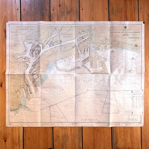 May include: A vintage nautical chart of the San Pedro Bay and Long Beach Harbors, folded and laid on a wooden surface. The chart features detailed illustrations of the coastline, waterways, and harbor infrastructure, with text and markings.