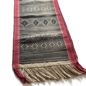 May include: A black and white woven table runner with a geometric pattern and red fringe. The runner is made of natural fibers and has a rustic look.
