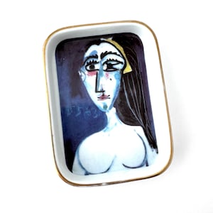 May include: A white ceramic rectangular dish with a gold rim. The dish features a painting of a woman's face with blue, yellow, and black details. The painting is signed "Picasso".