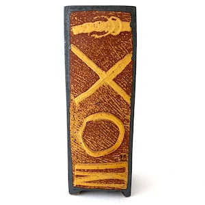 May include: A rectangular ceramic vase with a dark gray exterior and a textured brown center. The center features yellow abstract designs, including an X, an O, and a horizontal line. The vase has a small, square base.