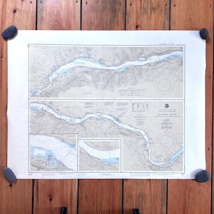 May include: A vintage nautical chart of the Columbia River, featuring detailed illustrations of the river's course and surrounding topography. The chart is printed in shades of blue and gray, and is held in place by four gray stones on a wooden background.