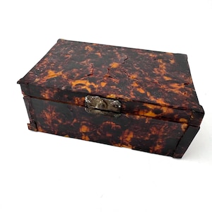 May include: A rectangular box featuring a dark brown and orange mottled tortoiseshell pattern with a cracked texture across the surface. The box includes a metallic latch mechanism on the front center for securing the lid to the base.