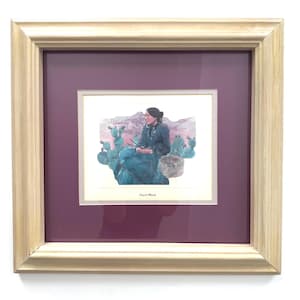 May include: Framed artwork featuring a seated figure in a desert landscape with cacti. The artwork is set against a deep purple mat and framed in a light-colored wood frame. The image has a vintage aesthetic.