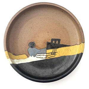 May include: A round ceramic plate with a brown rim and a central design. The design features a boat-like shape with yellow, black, and white stripes, and a figure. The plate has a speckled, earthy texture.