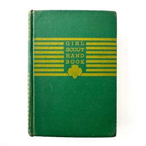 May include: A vintage Girl Scout Handbook with a green cover and gold lettering. The title "GIRL SCOUT HAND BOOK" is displayed prominently, along with a trefoil emblem. The book's cover features horizontal gold stripes.