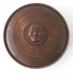 May include: A round, brown ceramic lid with a raised face design in the center. The lid has a textured surface and a darker brown rim. The face is detailed with a smiling expression.