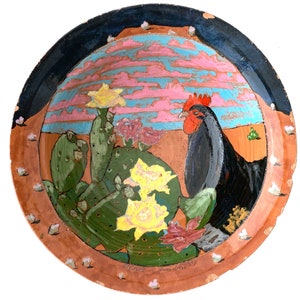 May include: A terracotta bowl with a painted design of a rooster, cactus, and flowers. The bowl has a brown rim and a white background with a blue sky and pink clouds.