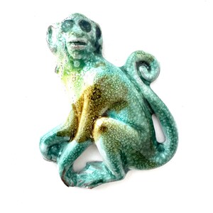 May include: A ceramic monkey figurine featuring a glossy, crackled glaze in shades of turquoise, teal, and olive green. The monkey is depicted in a seated position with its tail curled upward, showcasing a textured surface and detailed facial features.