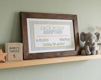May include: A framed cross-stitch birth announcement featuring the name Jack Noah Ashford, the date 6.19.2021, and the weight 8lbs 5oz, accented with green stripes and a heart. Beside it sits a small book titled Baby's Firsts and a gray plush elephant.