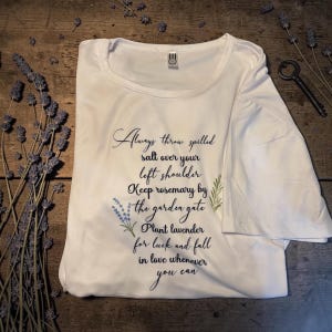 May include: White t-shirt with a round neck, featuring script text and botanical illustrations. The text reads "Always throw spilled salt over your left shoulder..." with lavender and rosemary accents. A key and lavender sprigs are on the wooden surface.