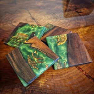 May include: Four square coasters made of dark wood with a green and gold resin inlay. The coasters have a unique, swirling pattern.