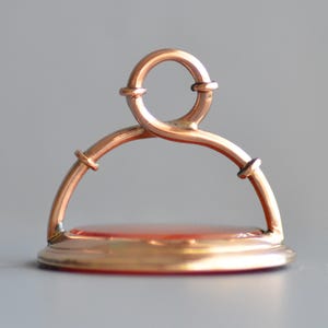 May include: A vintage gold-toned seal with a red stone base. The seal features a curved handle with a circular top and is designed with intricate details. The base is a flat, circular shape, and the overall design is elegant.