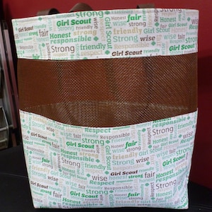May include: A brown mesh tote bag with a white fabric lining. The fabric lining has a repeating pattern of the words "Girl Scout" and words like "strong", "honest", "respect", "wise", "friendly", and "responsible" in green and brown text.