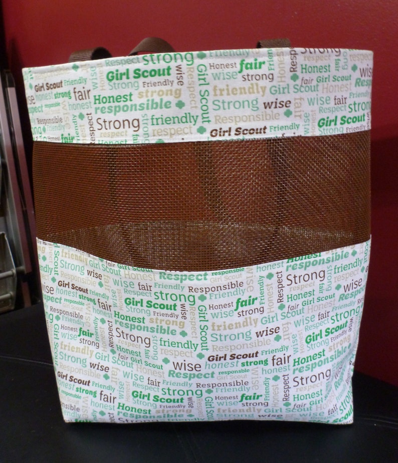 May include: A brown mesh tote bag with a white fabric lining. The fabric lining has a repeating pattern of the words "Girl Scout" and words like "strong", "honest", "respect", "wise", "friendly", and "responsible" in green and brown text.