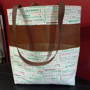 May include: A brown and white tote bag with a mesh pocket. The bag has a brown strap and is printed with the words "Girl Scout", "Strong", "Respect", "Honest", "Wise", "Friendly", and "Responsible" in green and brown lettering.