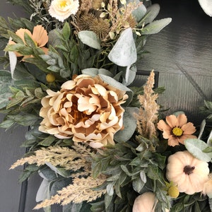 May include: A decorative wreath featuring beige peonies, small white pumpkins, and various dried grasses. The arrangement includes frosted green foliage, tan cosmos flowers, and spiky seed pods, creating a textured, layered botanical display.