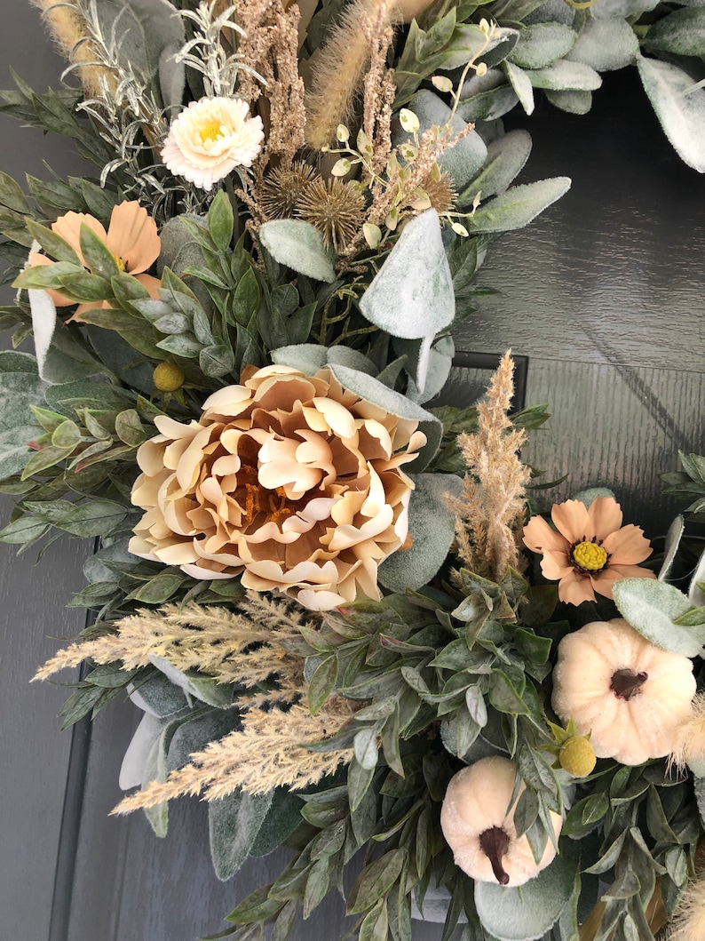 May include: A decorative wreath featuring beige peonies, small white pumpkins, and various dried grasses. The arrangement includes frosted green foliage, tan cosmos flowers, and spiky seed pods, creating a textured, layered botanical display.