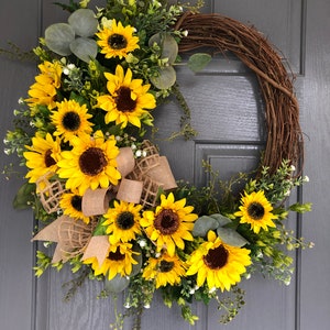 May include: A sunflower wreath with a burlap bow, featuring a mix of sunflowers and greenery. The wreath is made with grapevine and has a rustic look.