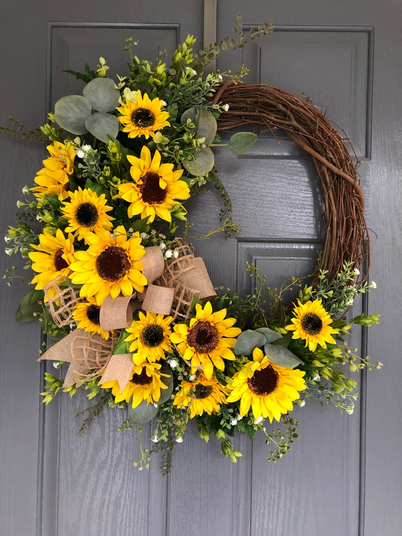 May include: A sunflower wreath with a burlap bow, featuring a mix of sunflowers and greenery. The wreath is made with grapevine and has a rustic look.