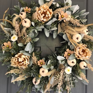 May include: A circular wreath featuring beige peonies, small white pumpkins, and light green lamb's ear leaves. The arrangement includes dried grasses, tan cosmos flowers, and spiky seed pods, all densely layered on a circular frame structure.
