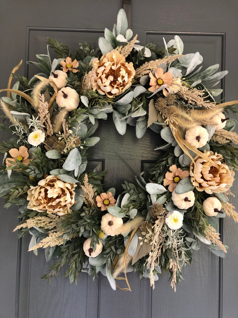 May include: A circular wreath featuring beige peonies, small white pumpkins, and light green lamb's ear leaves. The arrangement includes dried grasses, tan cosmos flowers, and spiky seed pods, all densely layered on a circular frame structure.