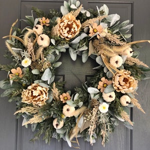 May include: A circular wreath featuring a dense arrangement of sage green lamb's ear leaves, dark green boxwood foliage, beige dried grasses, light peach-colored peony-style flowers, and small white pumpkins with dark stems.