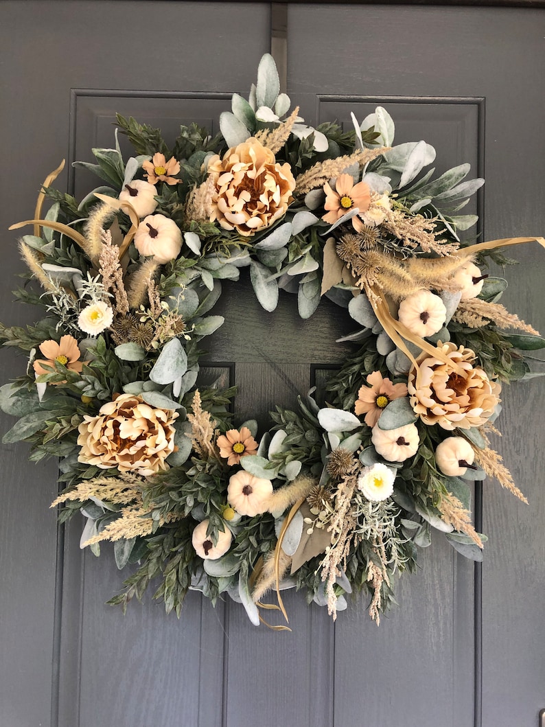 May include: A circular wreath featuring a dense arrangement of sage green lamb's ear leaves, dark green boxwood foliage, beige dried grasses, light peach-colored peony-style flowers, and small white pumpkins with dark stems.
