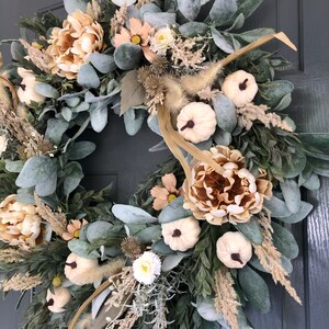 May include: A circular wreath featuring sage green lamb's ear leaves, beige peonies, small white pumpkins with dark stems, dried grasses, and thistle accents. The arrangement is densely packed with varied textures and neutral earth tones.