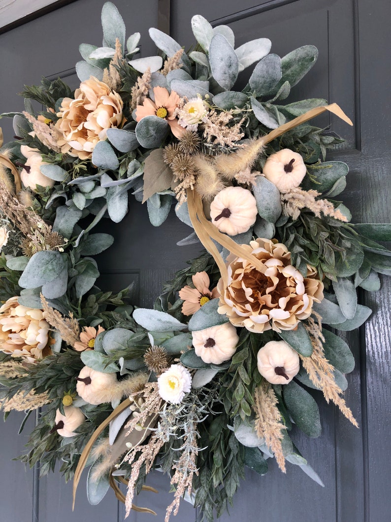 May include: A circular wreath featuring sage green lamb's ear leaves, beige peonies, small white pumpkins with dark stems, dried grasses, and thistle accents. The arrangement is densely packed with varied textures and neutral earth tones.