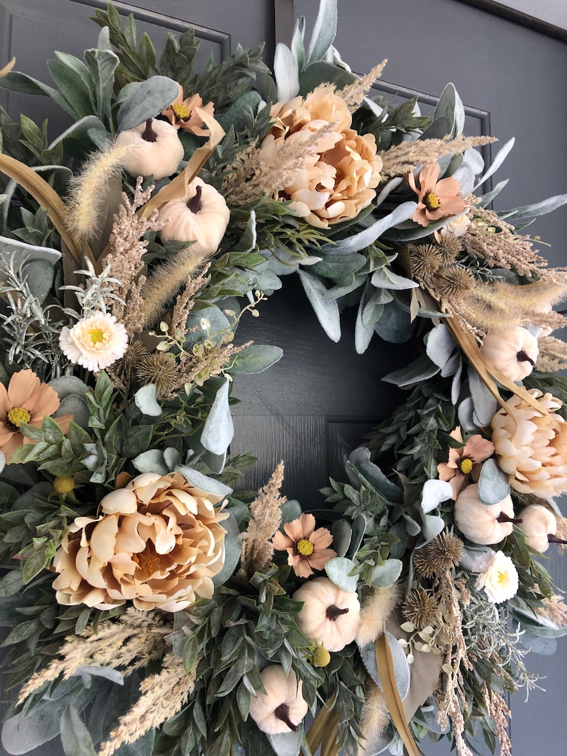 May include: A circular wreath featuring beige pumpkins, large peach-colored peony flowers, small cosmos blossoms, and dried grasses. The arrangement is accented with sage green lamb's ear leaves and dark green foliage, creating a textured display.