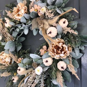 May include: A circular wreath featuring a mix of sage green lamb's ear leaves, beige peonies, small white pumpkins with dark stems, dried grasses, and spiky seed pods. The arrangement is dense and layered, creating a full, textured botanical display.