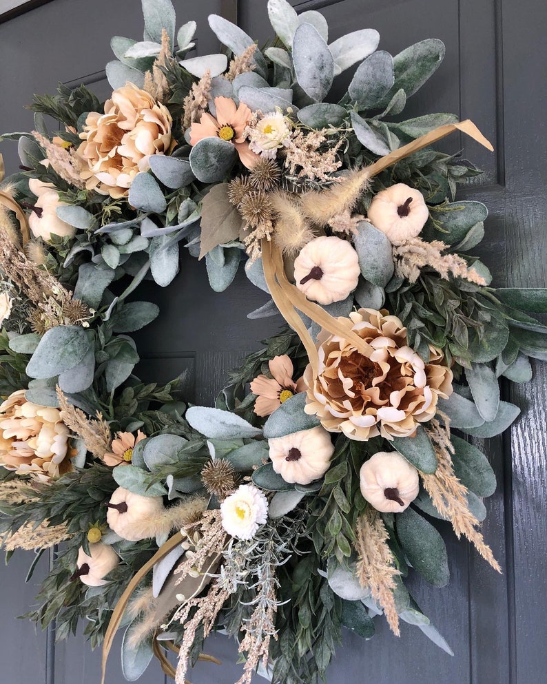 May include: A circular wreath featuring a mix of sage green lamb's ear leaves, beige peonies, small white pumpkins with dark stems, dried grasses, and spiky seed pods. The arrangement is dense and layered, creating a full, textured botanical display.
