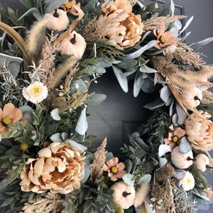 May include: A circular wreath featuring peach-colored peonies, small cream-colored pumpkins, and various dried grasses. The arrangement includes sage green lamb's ear leaves, textured brown seed pods, and small light-colored flowers on a dark base.