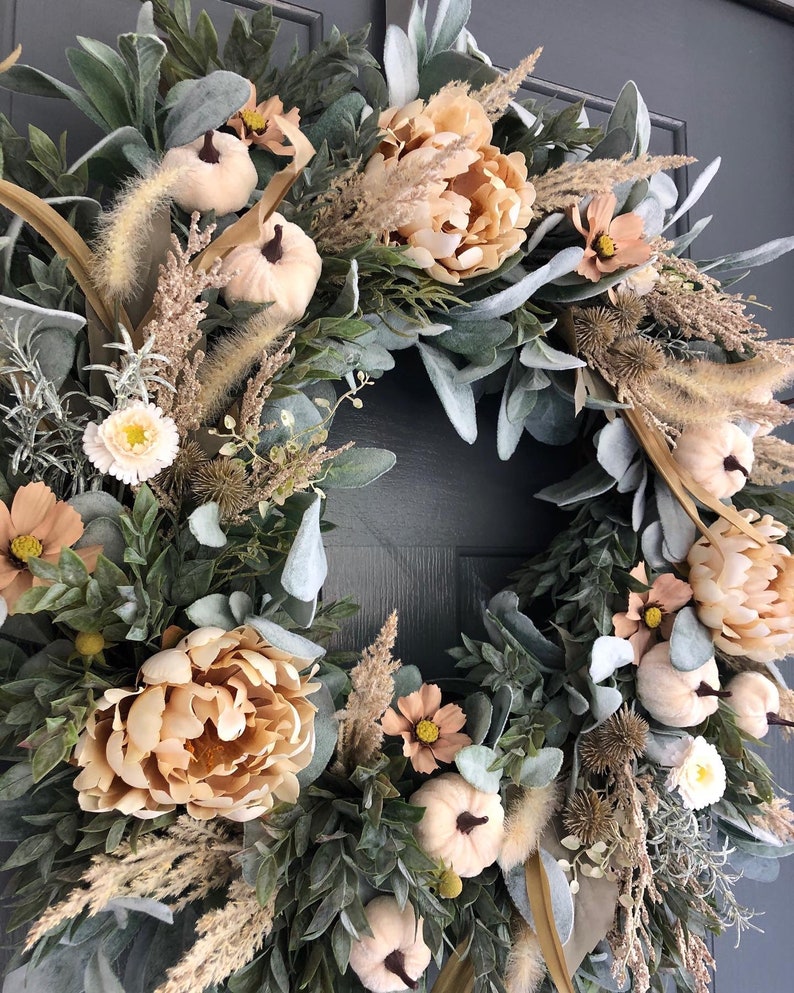 May include: A circular wreath featuring peach-colored peonies, small cream-colored pumpkins, and various dried grasses. The arrangement includes sage green lamb's ear leaves, textured brown seed pods, and small light-colored flowers on a dark base.