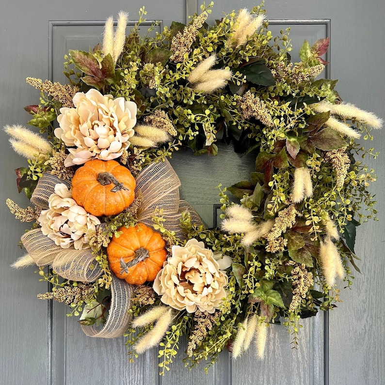 May include: A fall-themed wreath with a green base, beige flowers, and orange pumpkins. The wreath is decorated with beige and brown ribbon and white pampas grass.