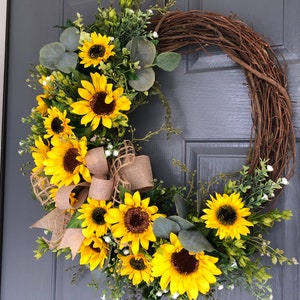 May include: A sunflower wreath with a burlap bow, featuring a mix of yellow sunflowers, green leaves, and white flowers. The wreath is hanging on a gray door.