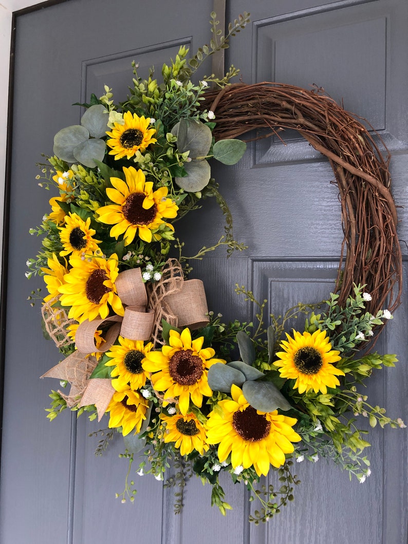 May include: A sunflower wreath with a burlap bow, featuring a mix of yellow sunflowers, green leaves, and white flowers. The wreath is hanging on a gray door.
