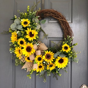 May include: A sunflower wreath with a burlap bow. The wreath is made of grapevine and features a variety of sunflowers in different sizes. The wreath is perfect for adding a touch of summer to your home decor.
