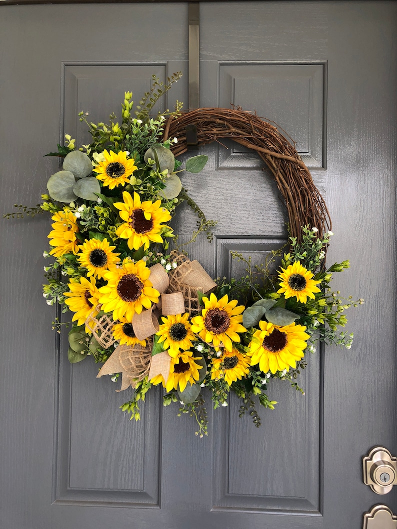 May include: A sunflower wreath with a burlap bow. The wreath is made of grapevine and features a variety of sunflowers in different sizes. The wreath is perfect for adding a touch of summer to your home decor.