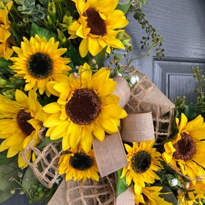 May include: A sunflower wreath with burlap ribbon and greenery. The wreath is decorated with multiple sunflowers and small white flowers.