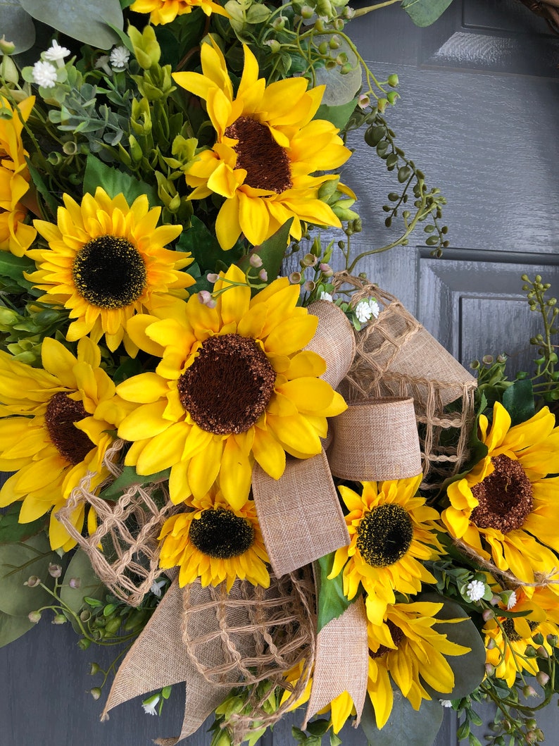May include: A sunflower wreath with burlap ribbon and greenery. The wreath is decorated with multiple sunflowers and small white flowers.
