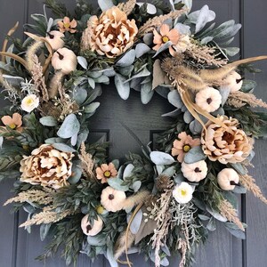 May include: A circular wreath featuring a mix of sage green lamb's ear leaves, beige peonies, small white pumpkins, dried grasses, and brown thistle accents. The arrangement is densely packed with various textures and neutral earth tones.