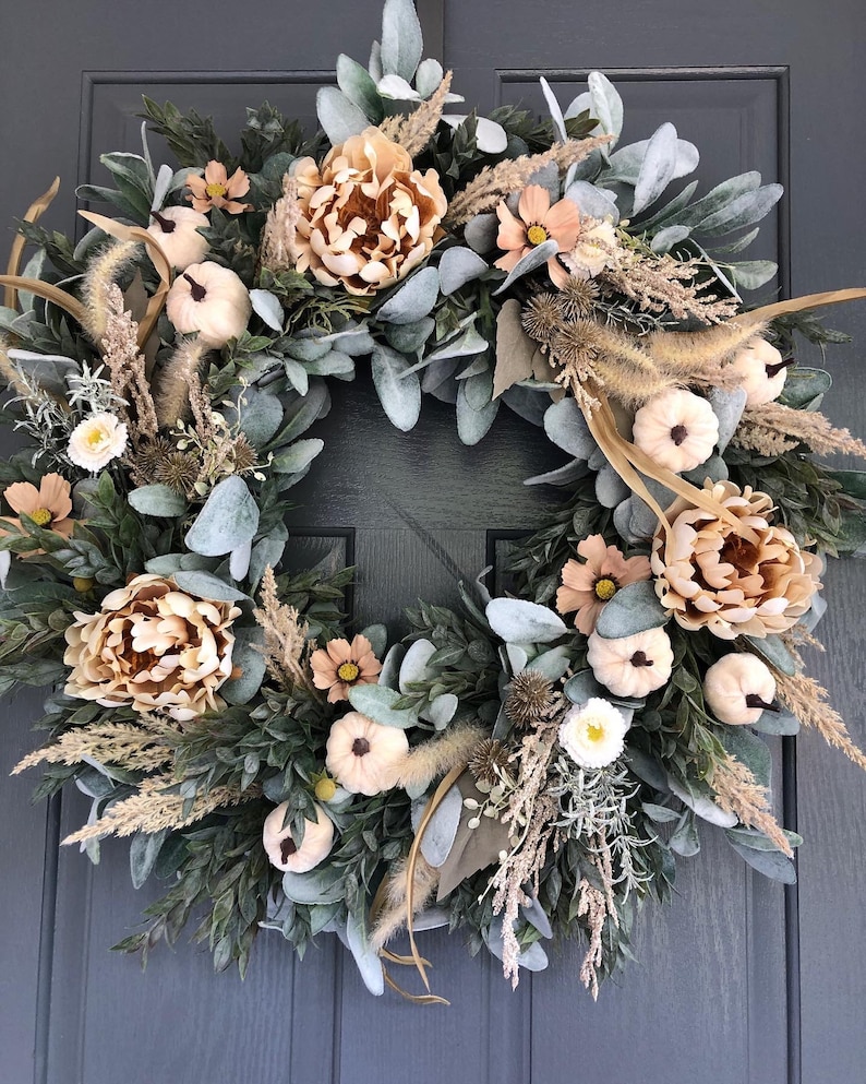 May include: A circular wreath featuring a mix of sage green lamb's ear leaves, beige peonies, small white pumpkins, dried grasses, and brown thistle accents. The arrangement is densely packed with various textures and neutral earth tones.