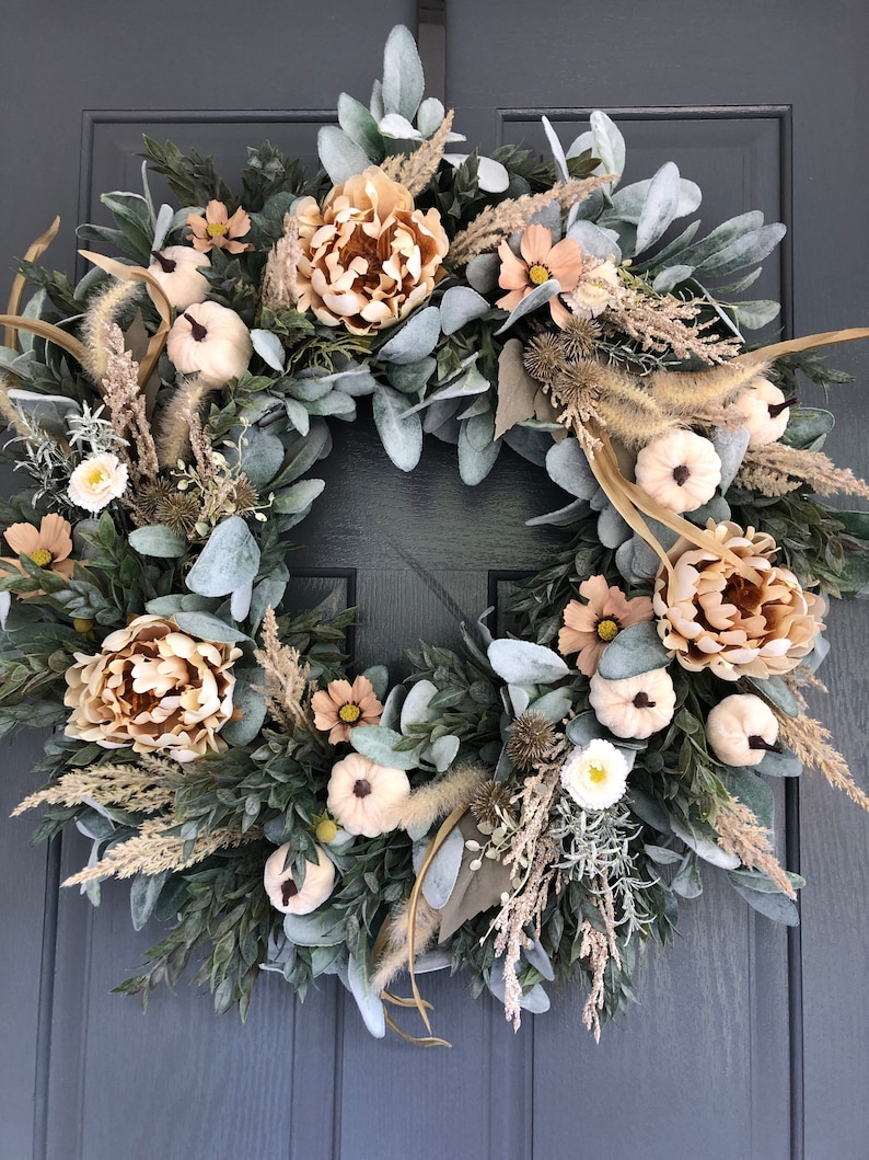 May include: A circular wreath featuring beige peonies, small white pumpkins, and light green lamb's ear leaves. The arrangement includes dried grasses, tan wildflowers, and dark green foliage, creating a textured, layered display of neutral tones.