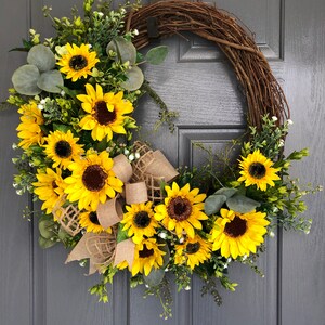 May include: A sunflower wreath with a burlap bow. The wreath is made of grapevine and features a variety of sunflowers, greenery, and small white flowers.